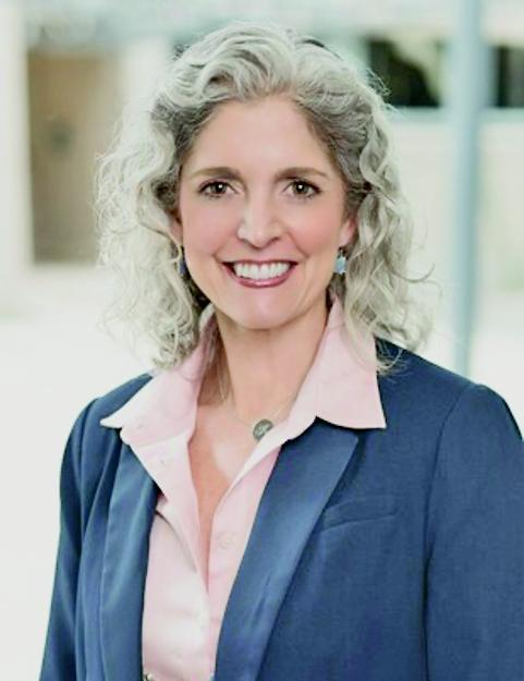 The image shows Dr. Karen Watts Barton, a professional woman with curly, shoulder-length gray hair. She is smiling warmly and wearing a navy blue blazer over a light pink blouse. A simple, elegant necklace adds a touch of detail to her outfit. The background is softly blurred, focusing attention on her confident and approachable demeanor.