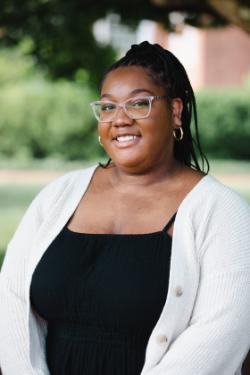 The image shows a person smiling warmly, wearing clear-framed glasses and hoop earrings. Their hair is styled in braids, and they are dressed in a black top paired with a light-colored cardigan. The background is outdoors, featuring greenery that is softly blurred, giving the portrait a pleasant and natural atmosphere.